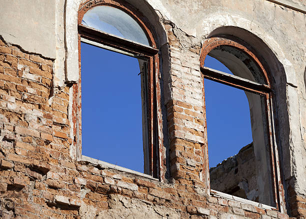 A pair of tall arched windows on a distressed building facade, featuring peeling paint, broken glass and exposed brickwork. The weathered textures and muted tones highlight the structure’s aged, decayed character. The scene conveys a sense of historic abandonment and rustic charm.