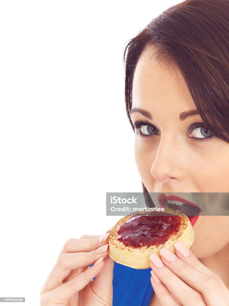 Young Woman Eating Toasted Crumpet and Jam Attractive young woman, with dark hair, eating a toasted crumpet with strawberry jam or jelly spread on top, looking towards the camera, wearing a blue top, isolated against a white background, clipping path, cut out 2015 Stock Photo