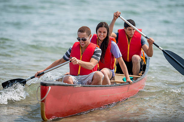 Young Teenagers on a Canoe Trip A multi-ethnic group of teenagers outside in a canoe on the water, wearing life jackets and paddling across the lake on a beautiful sunny day. canoe photos stock pictures, royalty-free photos & images