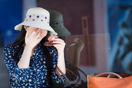 Pretty young woman holding a mirrir in her hand and looking at her face. She has long dark hair, she is wearing white straw hat with polkadots ribbon and dress with floral pattern Side view. An orange beach bag beside the woman. Reflection in the window behind. Outdoors, Europe. Nikon D800, full frame, XXXL. Blurred background. Minimal post-processing.