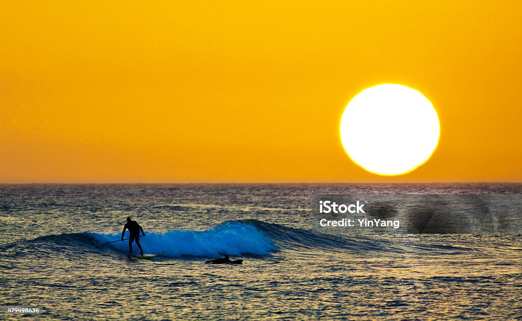 Sunset Surfer and Paddle Board on Pacific Waves, Kauai, Hawaii Surf board surfer and paddle board enthusiast surfing the waves and paddling a paddle board along the beach at sunset, in the Pacific Ocean waters off Poipu Beach, Kauai, Hawaii, USA. Horizontal seascape with unidentifiable silhouettes of people, with copy space. Hawaii Islands Stock Photo