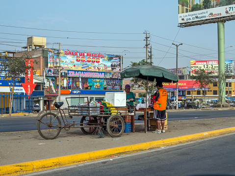 Lima, Peru - January 22, 2015: Street trader selling food from her mobile stall to man in reflective jacket. The stall is sited on the pavement between two lanes of road
