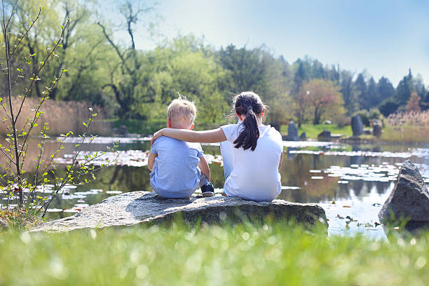 Two children, older sister and younger brother sitting on the shore of the lake.