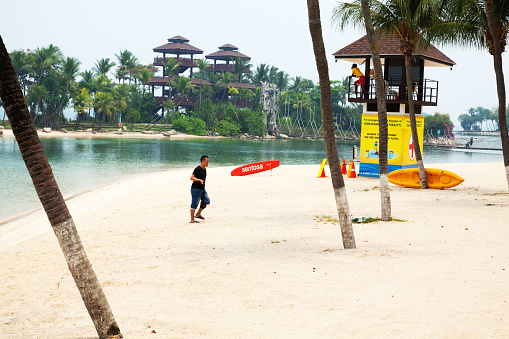 Singapore, Singapore - September 6, 2012: An asian man is walking in sand of beach on Sentosa Island. In background is tower of bay watch and coat guard. A man is standing on top. On beach is a red surf board with name Sentosa. In background is small island with wooden building and lookout.