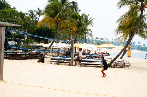 Singapore, Singapore - September 6, 2012: Scene on a beach of Sentosa Island at forenoon. A man is playing beach volleyball. In background are sun loungers and parasols amd people relaxing on beach. A man is opening parasol. Palm trees are on beach.