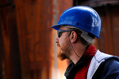 Young red head worker with protective helmet and sunglasses on a building site. Due to rusty background the worker can be seen also as a steelworker, or a oil rig worker. Nice crane and structure reflection on the helmet.