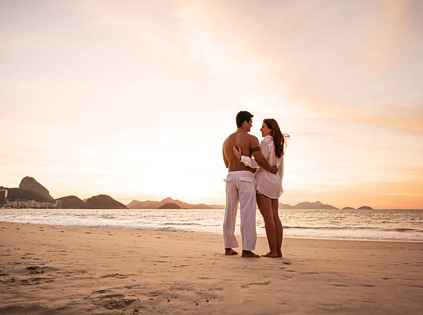 Vibrant Rio de Janeiro beach scene shows a lively shoreline bordered by a busy city road, tall buildings, and green hills, as turquoise waves roll onto golden sand under a clear blue sky.