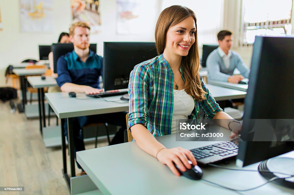 Young beautiful girl working on a computer Young beautiful girl working on a computer in a classroom with her classmates in the background Educational Exam Stock Photo