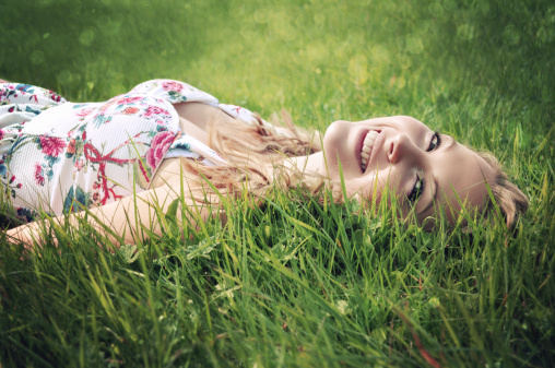 Young blonde woman with shoulder length hair and wearing a flower print summer dress lying down in the grass and smiling.