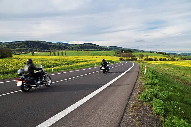 Photo of Motorcycles traveling along an empty road between blooming rape fields