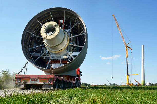 Semi-Truck transporting hubcap gear equipment of a wind turbine, slowly driving along a country road through green fields. Large mobile crane and tower of a new turbine in the background. Taken under clear sky at a construction site in the German North Sea Region, Lower Saxony, Germany, Europe.