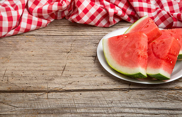 A weathered picnic table with a red and white checkered tablecloth and slices of seedless watermelon. The shot is from a higher angle leaving copy space. The image can be used to illustrate summer, food, summer food, fruit, farmers markets, summer food recipes, picnic, healthy summer food ideas, fourth of July, Labor Day, etc.