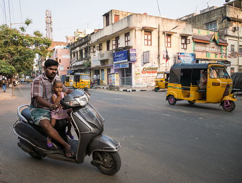 Chennai, India - 24 March 2015: Candid urban street scene on the hot streets of Chennai, India, with a father driving a moped with his daughter in front of him.  Tuk Tuks (motorised rickshaws) visible in the background.