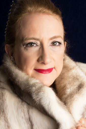 Vertical studio shot on dark blue of glamourous mature woman. Hair back, professional makeup and light fur coat. Smiling head and shoulders.