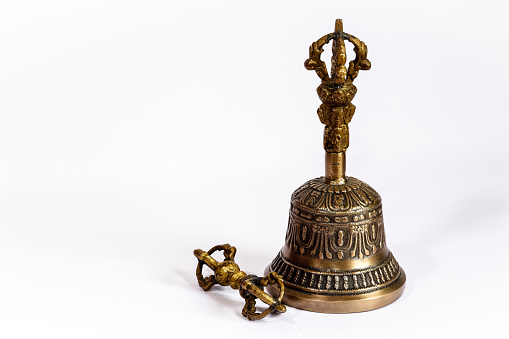Hand crafted, bronze Tibetan Buddhist Prayer Bell on a white background. The ringer by the side is either used to ring the bell or, to twirl around the base of the bell to make a continuous sound with the help of the pattern on the bell. A face of the Buddha can be seen on the handle.  Photo shot in studio environment; horizontal format.  Copy space.  Close-up.