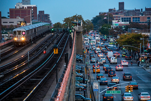 Red and green lights: NY Metro train on Line 7 elevated track in Queens speeds towards Manhattan at dusk. On the road below, 5 lanes of traffic try to exit the city in the opposite direction, their red brake lights on and going nowhere, despite the green traffic lights.  Station platform in centre frame.  Horizontal, HDR (light tone-mapping), High ISO with some noise evident, multiple logos, illegible vehicle plates, copy space.