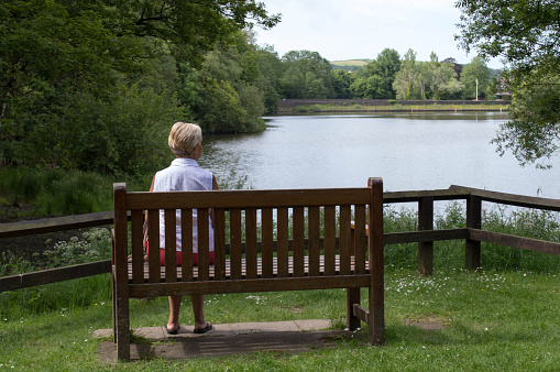 Image of a mature woman outdoors sitting on a bench looking over a lake in summer