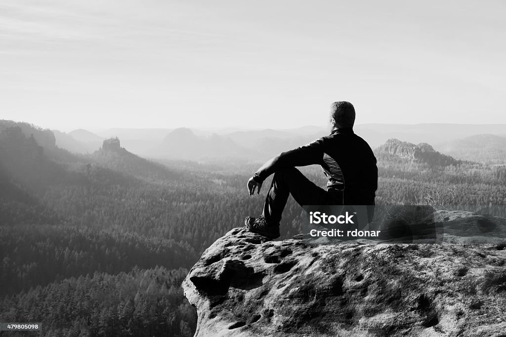 Tourist hiker man on the rock peak  in rocky mountains Young man in black sportswear is sitting on cliff's edge and looking to misty valley bellowHigh tourist hiker man on the rock peak  in rocky mountains 2015 Stock Photo