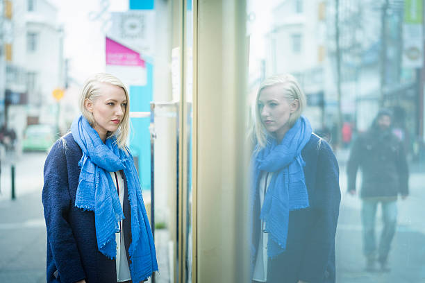 A photo of young female looking at store window. Blond woman is looking at display. She is in warm clothes. Reflection of her is falling on glass.