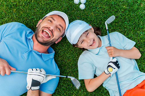 Top view of cheerful little boy and his father holding golf clubs and smiling while lying on the green grass
