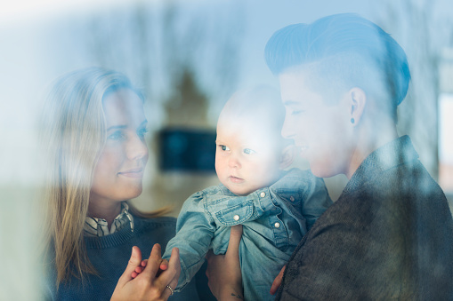 Loving lesbian couple with baby at home. Happy female partners with cute toddler in house. Homosexual couple and kid are seen through transparent glass window.