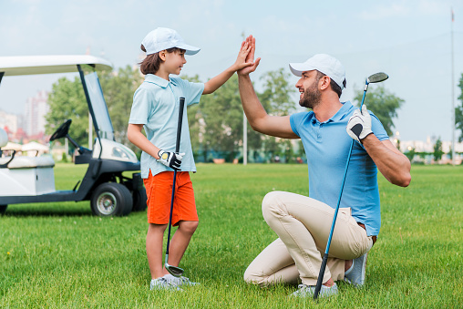 Cheerful young man and his son giving high-five to each other while standing on the golf course