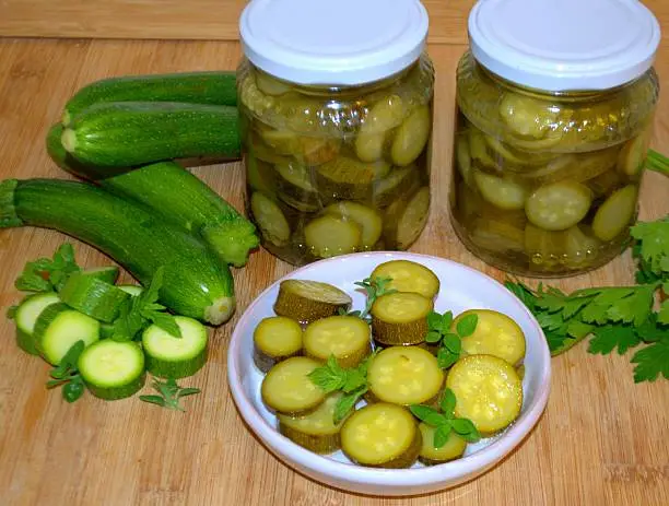 In the foreground flat, round white with round slices of zucchini, with small mint leaves; on the right some green leaves of parsley; on the left four green zucchini and 6 round slices of zucchini with small mint leaves; at the bottom, two jars with a white lid, with green zucchini in sweet and sour liquid.