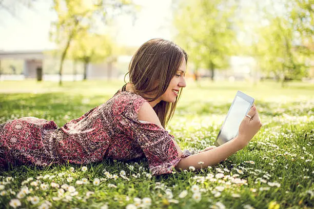 Profile of young smiling woman enjoying in grass during spring day and using touchpad.