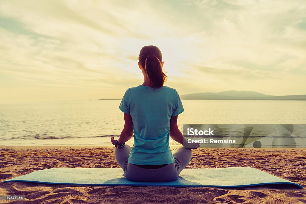 Girl meditating in pose of lotus on beach Young woman meditating in pose of lotus on beach near the sea at sunset in summer, rear view 2015 Stock Photo