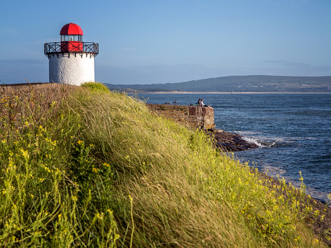 Burry Port, UK - July 4, 2015: Two cyclists sitting on sea wall relaxing alongside white lighthouse and Welsh coast
