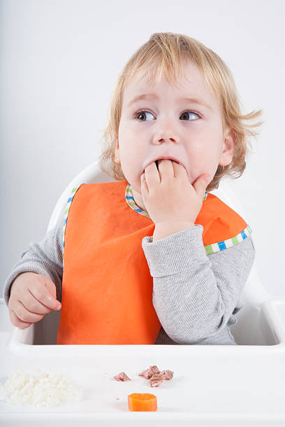 baby eating hand blonde caucasian baby seventeen month age orange bib grey sweater eating meal in white high-chair surprised looking at carrot and tuna kids eating tuna stock pictures, royalty-free photos & images