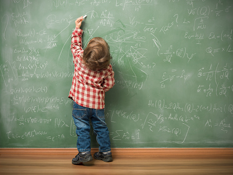 Little boy writing on green blackboard with math formulas written on.He is wearing a red plaid shirt and standing on the left side of frame.The green board is full of mathematics and physics formulas.He is seen in full length and holding chalk in left hand.The photo was shot in studio with a medium format camera Hasselblad H4D.