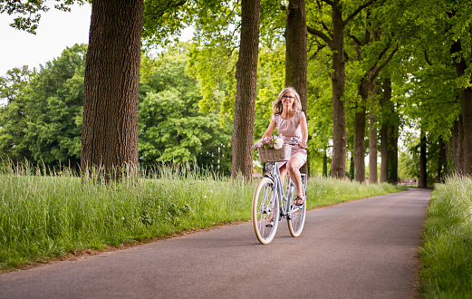 Full length shot of a mature woman cycling past trees along a country road through a lush summer park