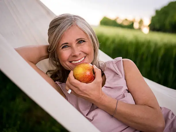 Healthy mature woman relaxing in a hammock in a park and eating a fresh apple