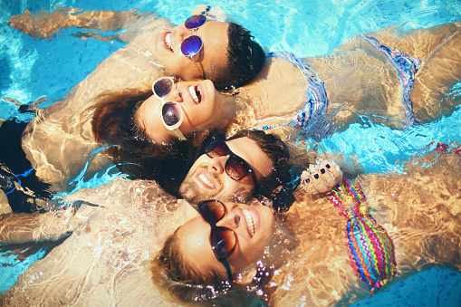 Top view of group of young adults relaxing in swimming on hot summer day. They are floating on water side by side, wearing sunglasses and facing the Sun. There are two guys and two girls, all smiling.