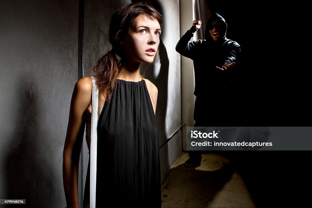 Woman In Danger at a Dark Alley Photo of a criminal stalking a woman alone in a dark street alley.  The thug is hiding in the shadows and the woman is alone and vulnerable in the street.  The criminal is a robber.  She is a scared victim. The image depicts crime and fear. 2015 Stock Photo