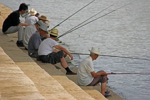 Pyongyang, North Korea  - August 15, 2013: A group of elderly men relax while fishing in the Taedong River. Here they are grouped together in the shade of the Taedong bridge on a hot morning in August.
