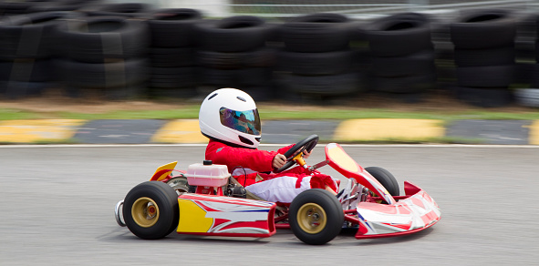 Asian boy wearing red and white race suit and white helmet practising in a gokart. Shot with a Canon 1DmkIV with slow shutter speed to create a panning speed blur effect with rubber tires in the background.