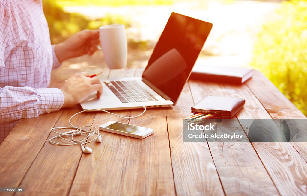 Businessman working outdoor Smart casual dressed person working on computer drinking coffee mug sitting at rough natural wooden desk outdoor with green tree and sun on background 2015 Stock Photo