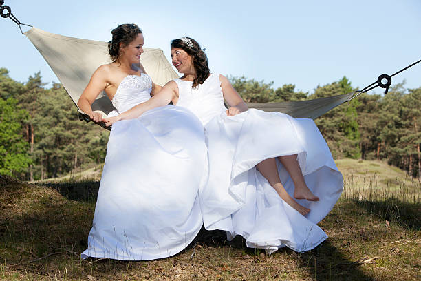 two brides look at each other in hammock against blue sky with forest background