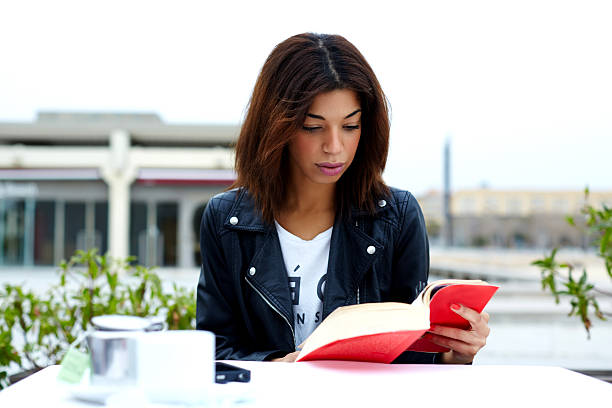 Attractive woman reading a book while sitting in a cafe Portrait of young lovely female enjoying a good book while sitting at the table in coffee shop terrace, charming afro american woman reading novel or book during her recreation time at weekend desiderius erasmus stock pictures, royalty-free photos & images