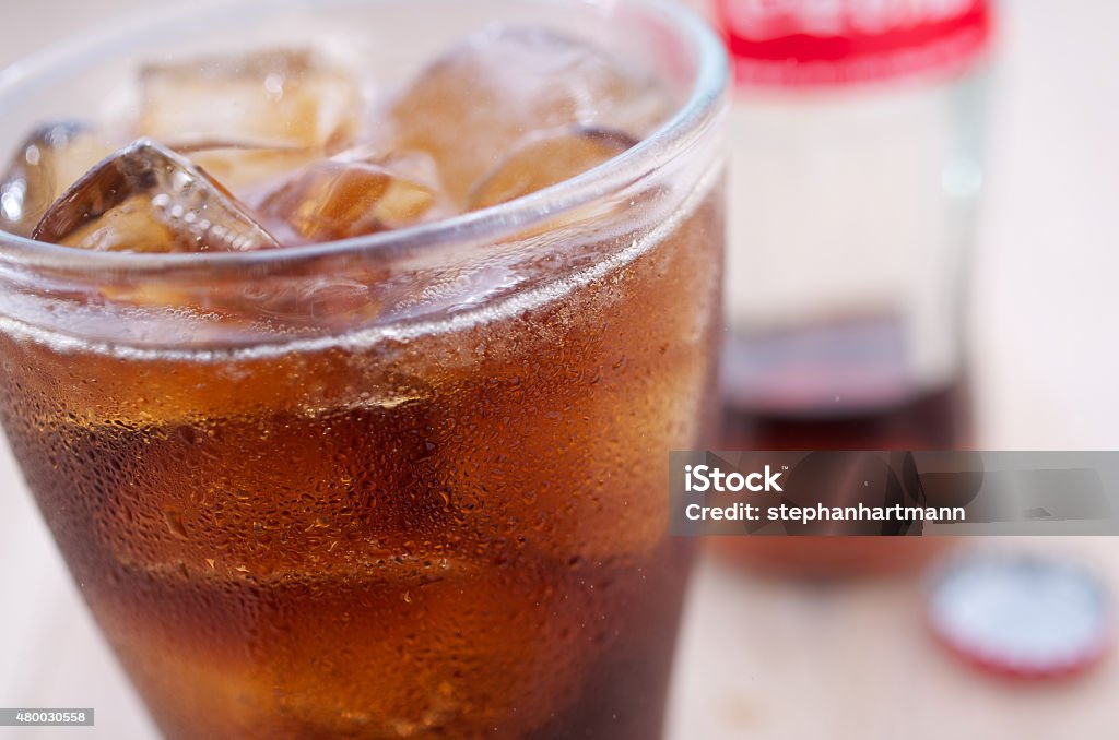 A glass of cold coke A glass of refreshing ice-cold cola with ice cubes. The cold glass has condensed water pots. In the background is a bottle on a wooden table.  2015 Stock Photo