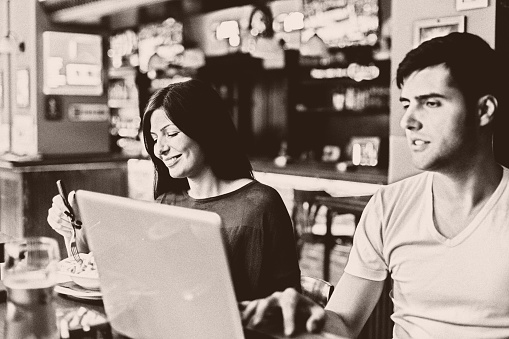 He working on laptop, she eating her pasta at restaurant