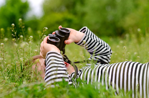 Young child using a pair of binoculars to look up into the sky as he lies on back in a grassy rural meadow enjoying a day in nature
