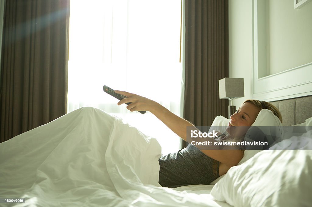 Cheerful girl watching TV from bed Cheerful young woman waking up in her hotel room watching tv. Bedroom Stock Photo