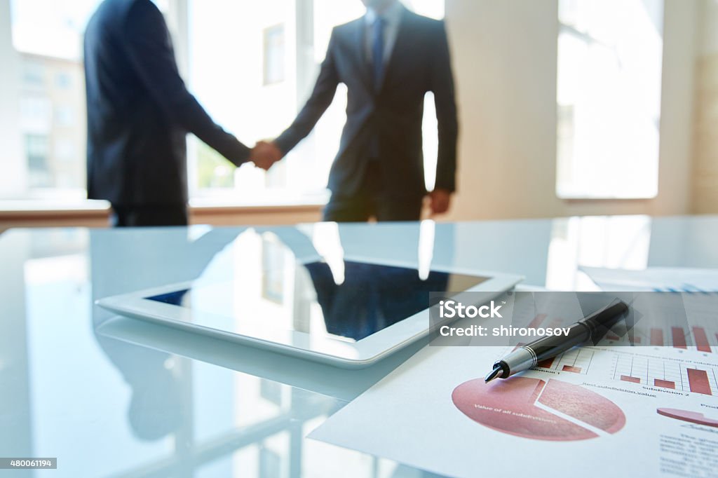 Touchpad, document and pen Business objects at workplace with handshaking partners on background 2015 Stock Photo