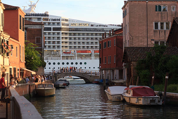 Venice, Italy - May 17, 2015: A huge cruise ship belonging to the Swiss-based MSC Cruises entering Venice on the Giudecca Canal at sunset, as seen from a small canal in the Dorsoduro area of Venice, Italy. Groups of people can be seen canal-side, on a bridge, and on the decks of the boat. Allowing large ships in the delicate Venetian lagoon has elicited controversy and protest.