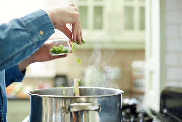 Midsection cropped image of woman adding cilantro to cooking pot. Focus is on her hand sprinkling healthy herb. She is preparing food in domestic kitchen.