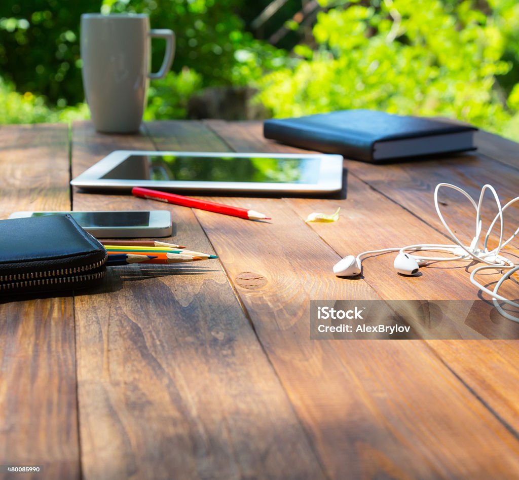 Modern lifestyle background Country rough wooden desk and lifestyle hobby items dropped around electronic devises pencils coffee mug notepad green flora and tree shadows behind Note Pad Stock Photo