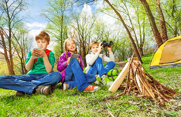Happy three children resting together near the wooden bonfire and yellow tent during camping in summer weather